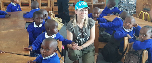 Eine Frau sitzt inmitten einer Gruppe von Kindern in einem Klassenzimmer. Die Kinder tragen blaue Schuluniformen.