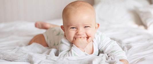 Ein lächelndes Baby liegt auf dem Bauch und steckt seine Hand in den Mund, während es auf einem Bett liegt.