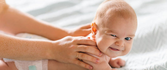 Ein Baby liegt auf dem Bauch und wird sanft massiert, während es schelmisch in die Kamera lächelt.