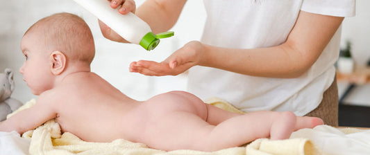 Eine Person trägt eine Lotion auf den Rücken eines Babys auf, das auf dem Bauch auf einem Handtuch liegt.
