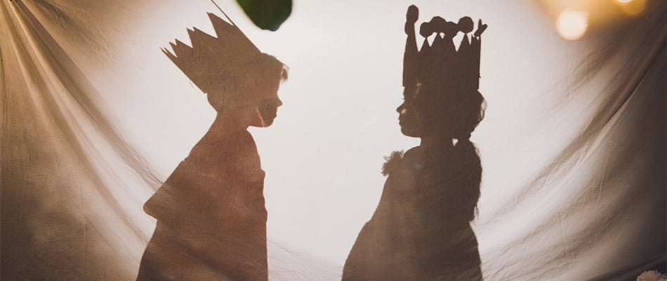 Zwei Kinder spielen verkleidet als Könige hinter einem Vorhang, ihre Schatten projizieren majestätische Kronen.