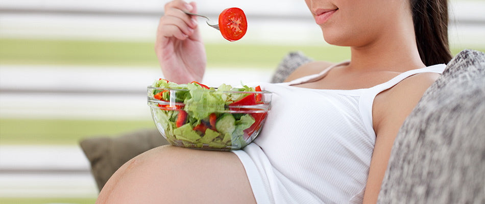 Schwangere Frau isst Salat mit einer Tomate auf einer Gabel, die Schüssel auf ihrem Babybauch.