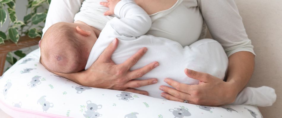 Eine Frau stillt ein Baby, das auf einem Stillkissen liegt. Beide tragen weiße Kleidung, und im Hintergrund sind Pflanzen zu sehen.
