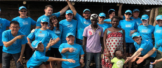 Eine Gruppe von Menschen in blauen T-Shirts und Hüten steht zusammen mit lokalen Bewohnern in Kenia vor einem Gebäude