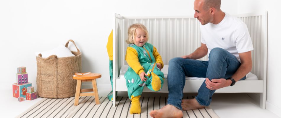 Ein blondes Mädchen im grünen Schlafsack mit Füßen sitzt auf dem Bett zusammen mit seinem Vater.