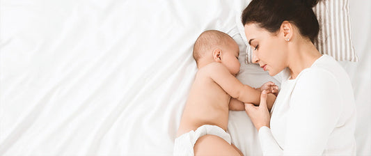 Eine Mutter liegt im Bett und hält sanft die Hand ihres schlafenden Babys, während beide sich aneinander kuscheln.