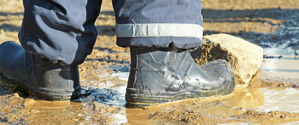 Ein Kind trägt Gummistiefel und steht in einer schlammigen Pfütze auf einem Feldweg.
