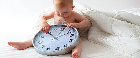 Ein Baby sitzt auf einem Bett und spielt mit einer großen Uhr. Es untersucht neugierig die Zeiger der Uhr.