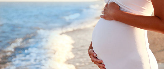 Eine schwangere Frau steht am Strand und hält ihren Bauch mit beiden Händen. Sie trägt ein weißes Kleid, und im Hintergrund sind das Meer und die Wellen zu sehen.