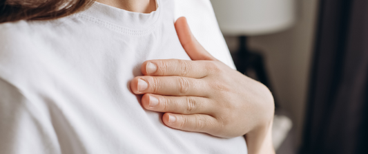 Eine Frau hält sich die Hand auf die Brust und trägt ein weißes T-Shirt. Im Hintergrund ist unscharf ein Lampenschirm zu sehen.