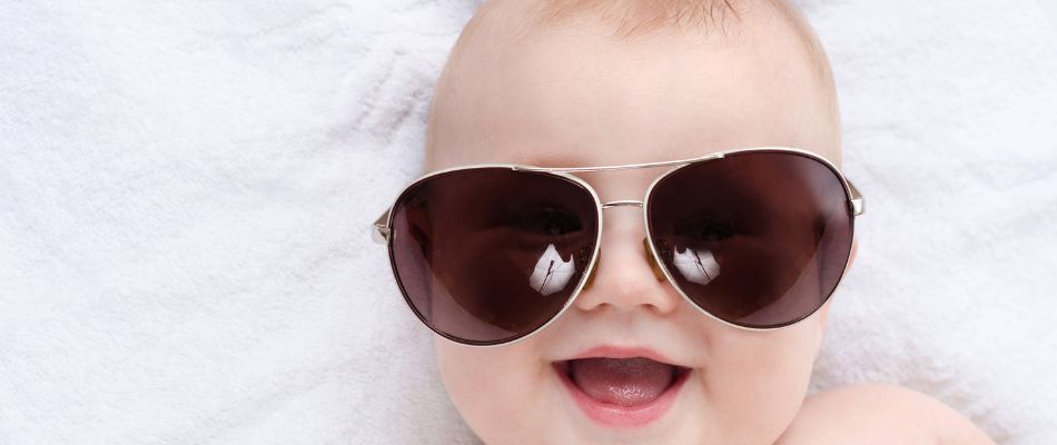 Ein fröhliches Baby liegt auf einer weißen Decke und trägt eine Sonnenbrille. Es lächelt breit.