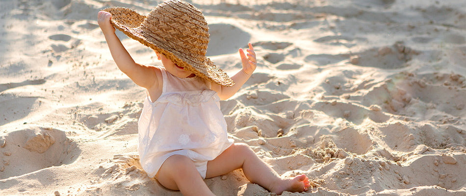 Ein kleines Kind sitzt im Sand an einem Strand und trägt ein weißes, ärmelloses Kleidchen. Es hält einen großen Strohhut auf dem Kopf und spielt im Sand.