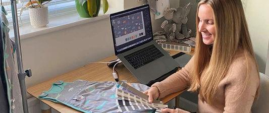 Designerin sitzt an einem Schreibtisch mit Laptop, Stoffproben und Babybekleidung, lächelnd bei der Arbeit.