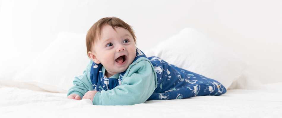 Ein fröhliches Baby liegt auf dem Bauch in einem blauen Schlafsack mit weißen  Mustern.