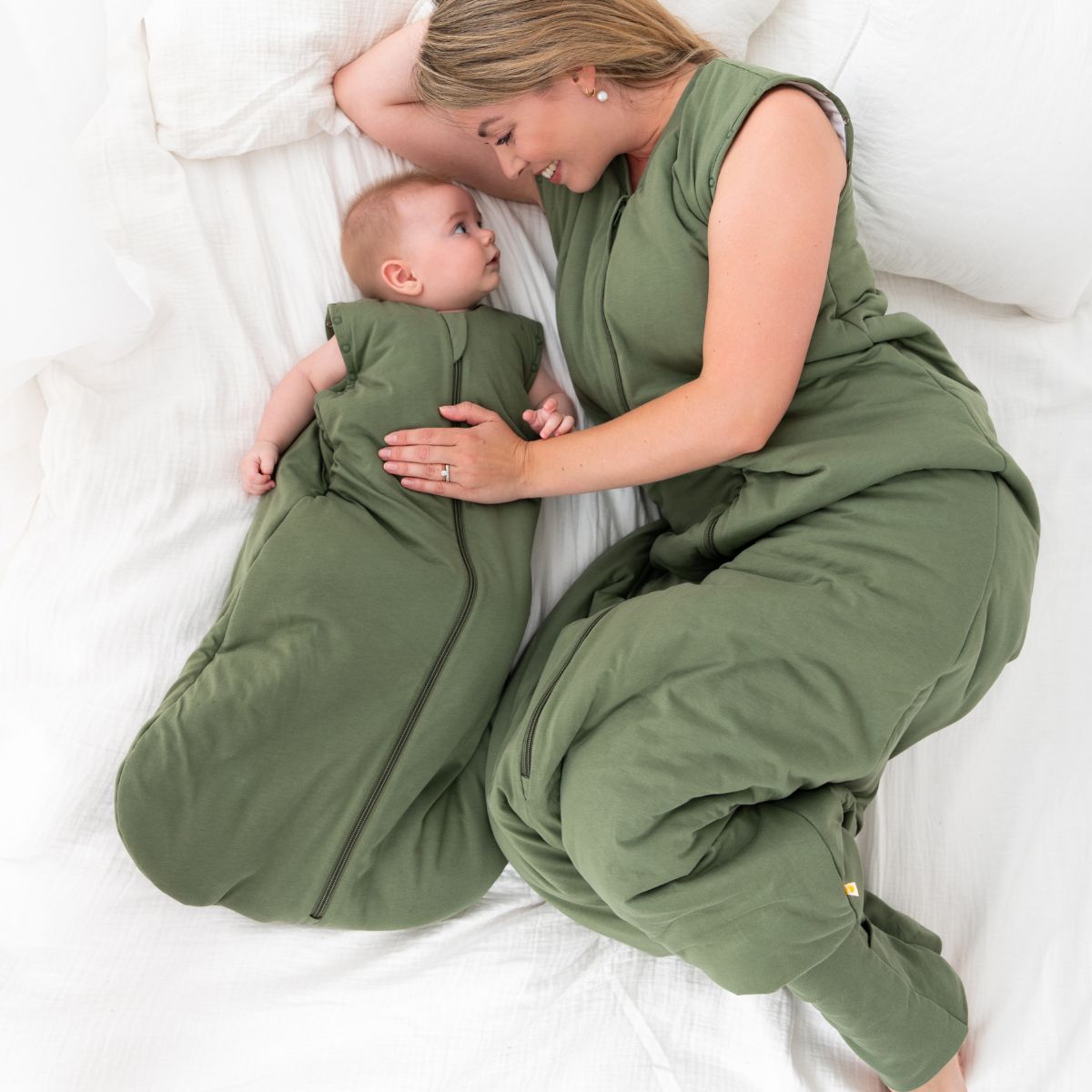 Eine Mutter liegt entspannt auf einem weißen Bett, gekleidet in einem grünen Schlafsack. Neben ihr liegt ihr Baby ebenfalls in einem passenden grünen Schlafsack. Beide schauen lächelnd.
